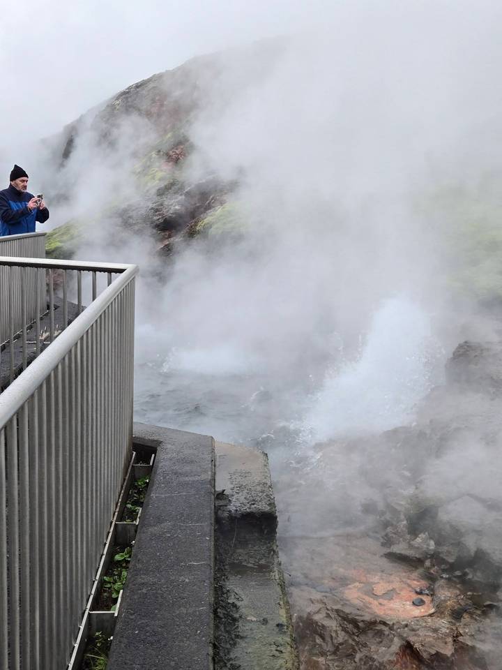 Billowing steam and boiling water surge from a geothermal vent behind safety railings.