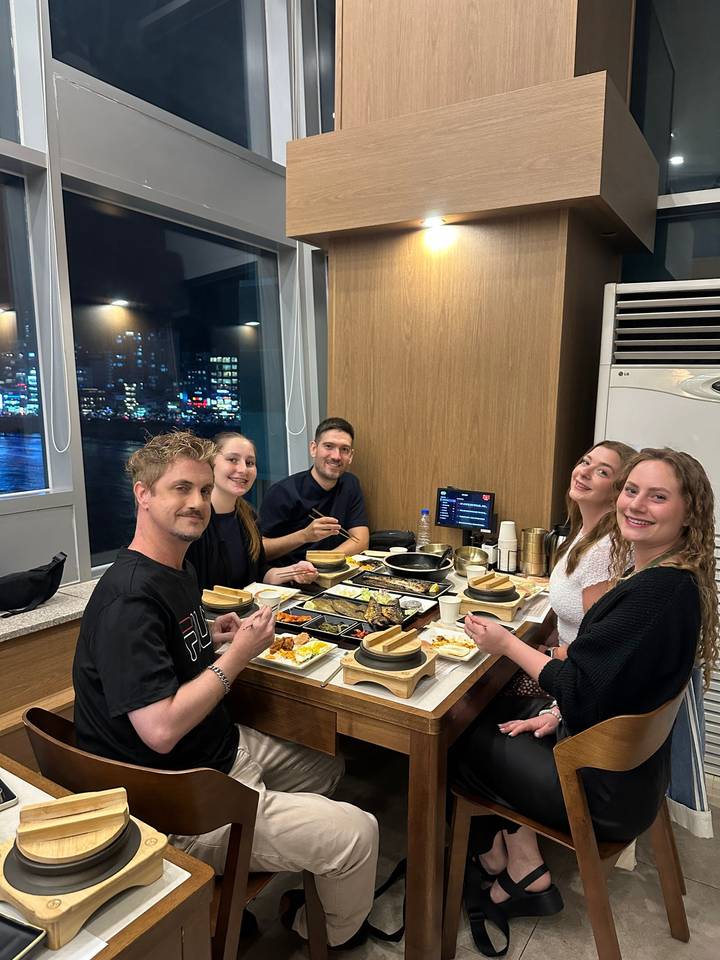 Five friends enjoy a Korean barbecue dinner at a modern restaurant table with a night-time city skyline visible through large windows behind them.