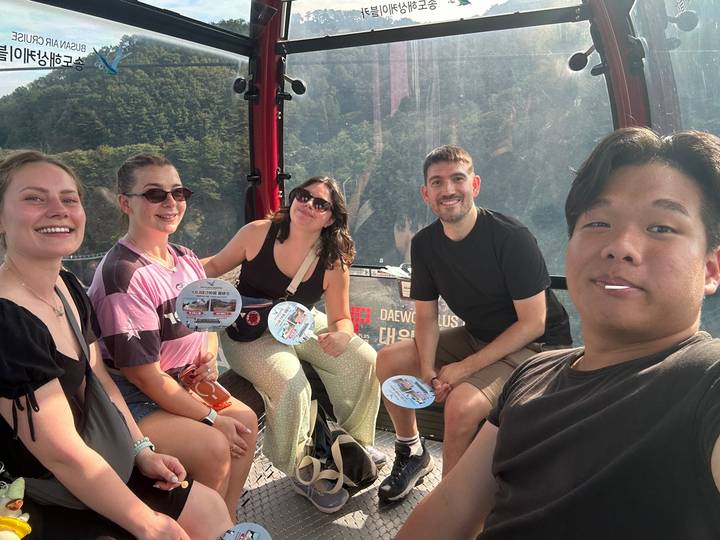 A cheerful group of young travellers rides inside a red cable car gondola with forested mountains visible through the glass.