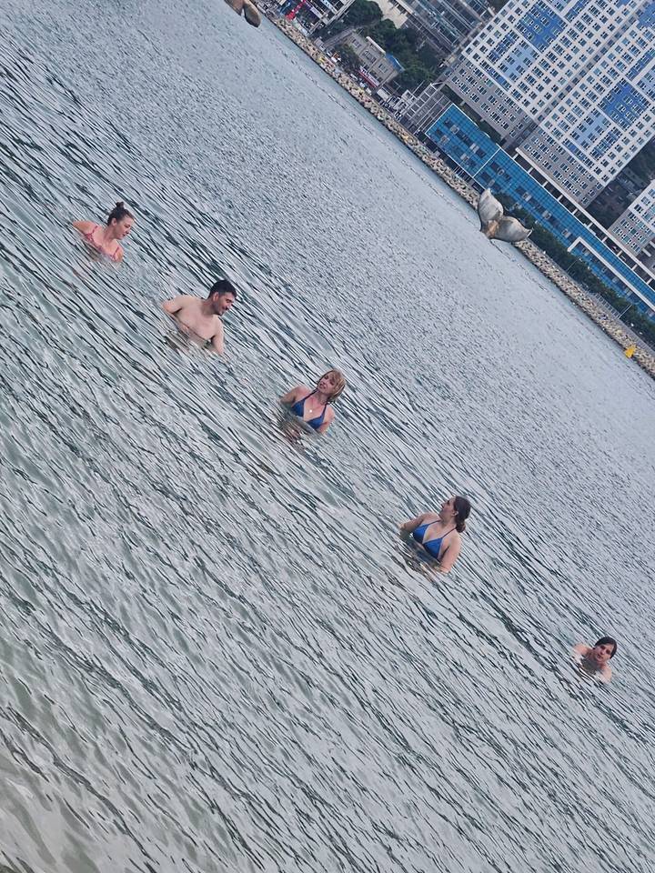 Three travellers in swimwear wade and chat together in calm coastal water near a stone shoreline.