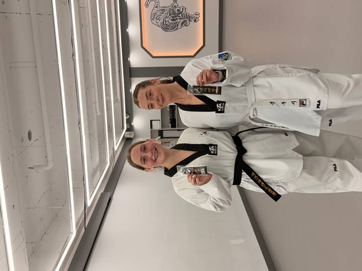 Two women proudly display their new black belts while dressed in crisp white taekwondo uniforms inside a bright martial-arts studio.