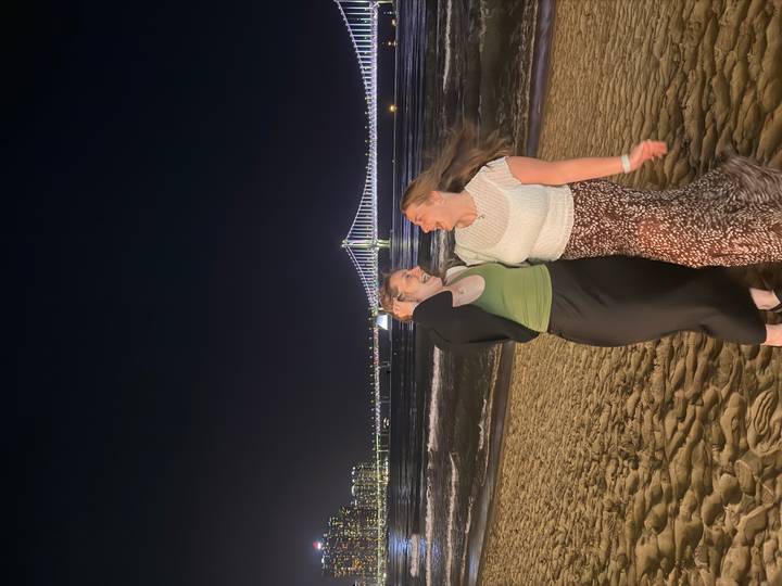 Two laughing friends pose on a sandy beach at night with Busan’s brightly lit Gwangan Bridge stretching across the dark sea behind them.