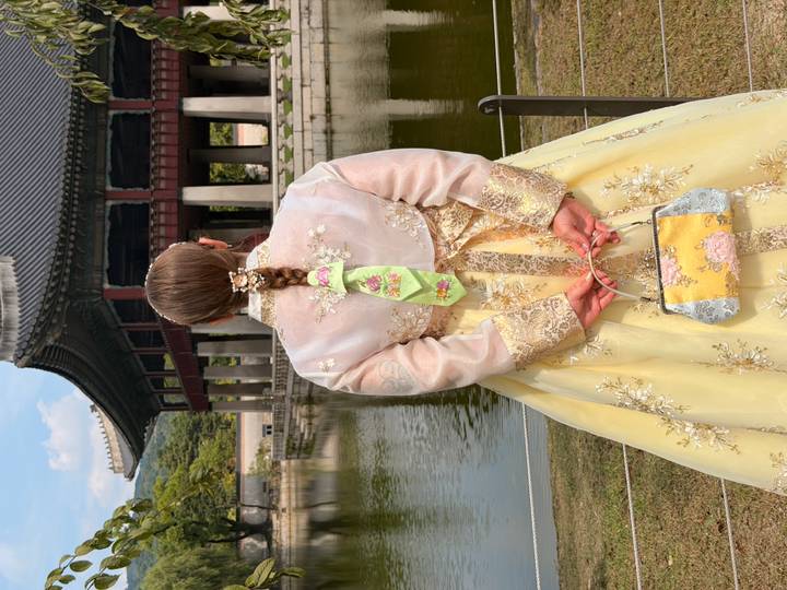 A woman in an elegant yellow hanbok stands beside a serene palace pond, ornate pavilion and tiled roof rising ahead of lush hills.