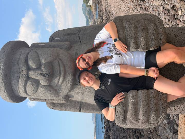 Two travellers smile while leaning against a large stone Dolhareubang statue on a rocky Jeju-do coast under a bright sky.