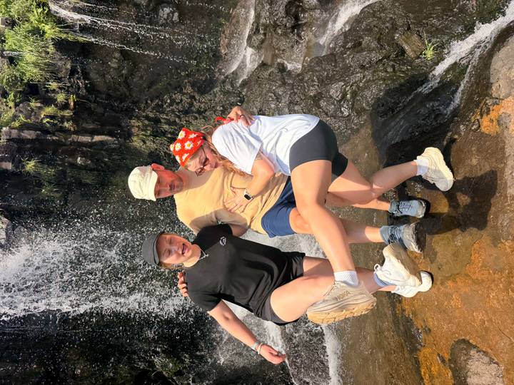 Three travellers laugh while balancing on a rock ledge beneath a cascading waterfall, splashes sparkling in bright sunlight.