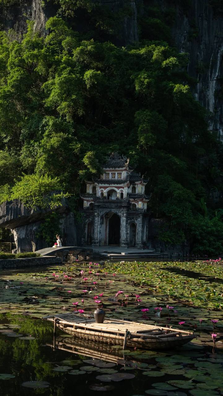 Ancient Vietnamese temple gate nestled in dense jungle cliffs with lone visitor in bright dress.
