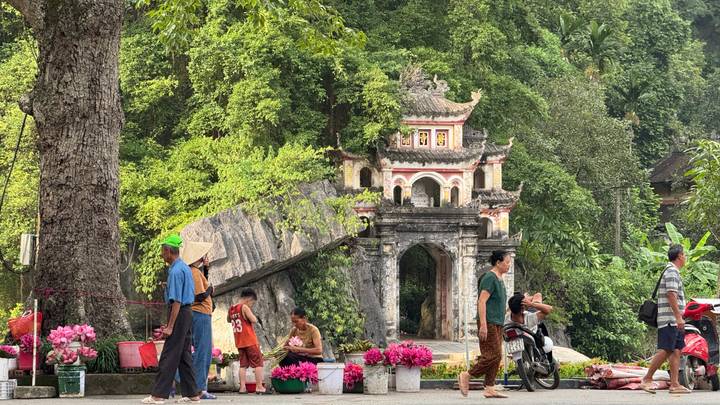 Local vendors sell pink lotus flowers near ornate jungle temple entrance in Vietnam.