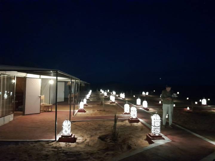 Nighttime desert camp walkway lined with glowing lanterns and canvas tents under starless sky.
