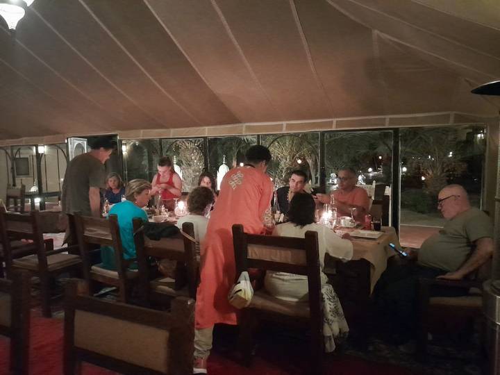 A large group of travellers sit around a long wooden table inside a canvas-roofed desert camp restaurant, dining by candlelight.