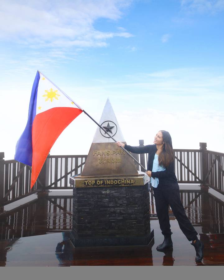 A smiling woman waves a national flag beside the Fansipan summit marker above the clouds.
