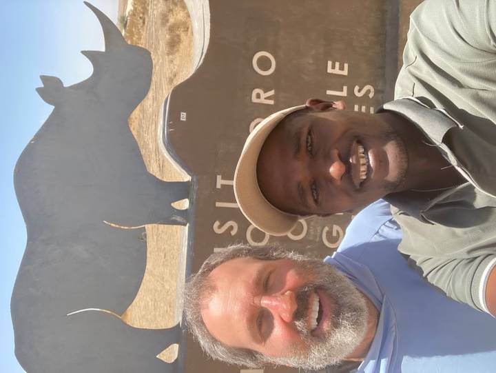Two smiling men taking selfie in front of sculpted rhino sign at reserve entrance.