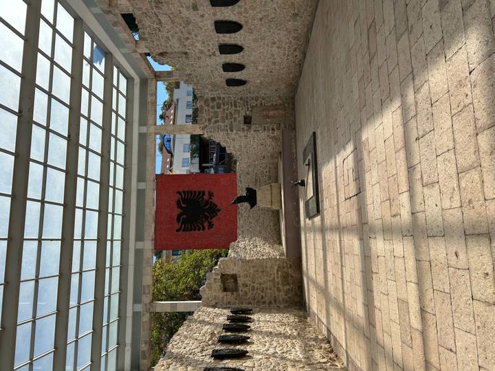 Open-air memorial with Albanian double-headed eagle mosaic flanked by ancient stone walls.