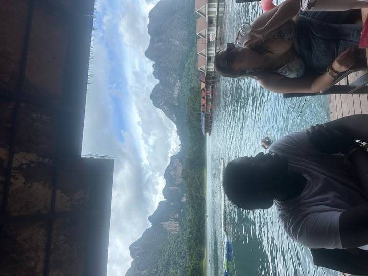 Travellers sit under a shaded roof overlooking a tranquil lake and karst peaks.