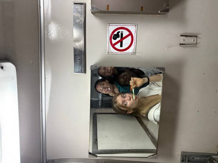 Friends jokingly brush teeth together in a small train washroom mirror.