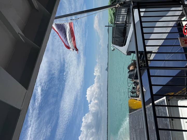 A Thai flag flutters from a ferry railing over turquoise sea beneath a bright sky.