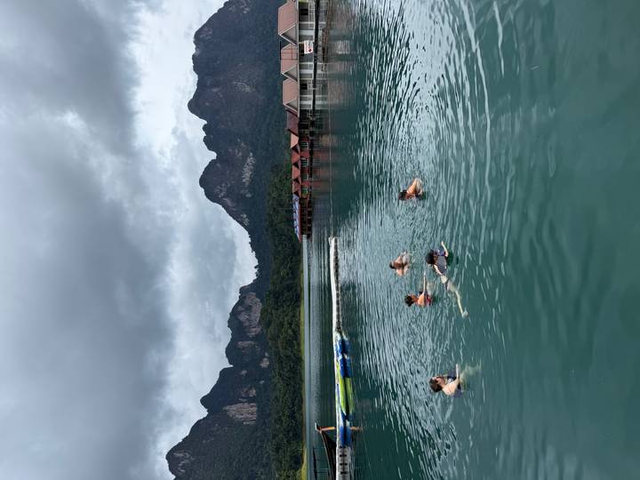 Travelers swimming in a calm emerald lake bordered by dramatic limestone mountains and floating bungalows.