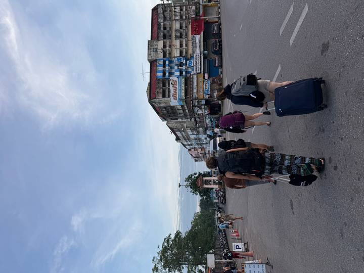 Backpackers pulling luggage through an urban Thai street lined with low-rise shop buildings under blue sky.
