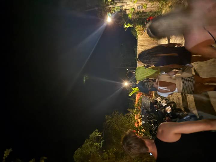 Young travelers walk down a dimly lit sandy path at night with motorbikes parked beside tropical foliage.