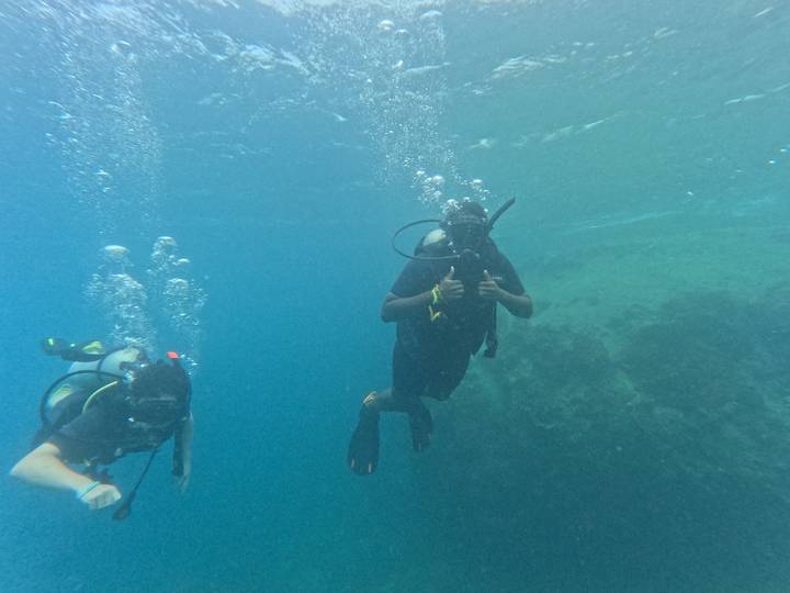 Two scuba divers give a thumbs-up while hovering over clear blue water next to a coral wall.
