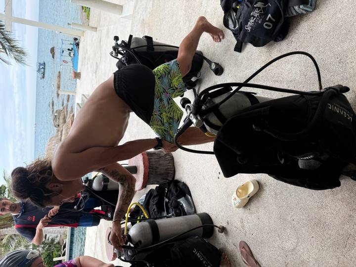 Diver in board shorts assembles scuba gear on shore with the sea and dive boats in the background.