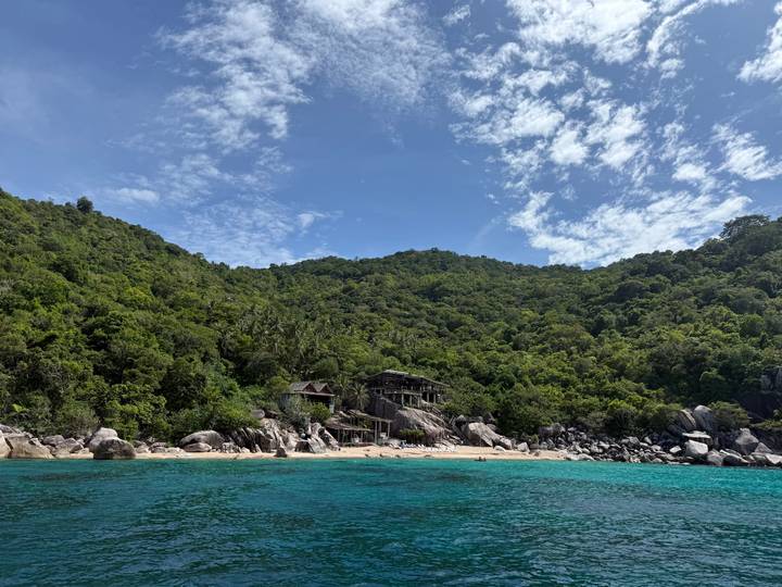 Lush green hillside meets turquoise sea and a secluded sandy beach under a bright blue sky with wispy clouds.