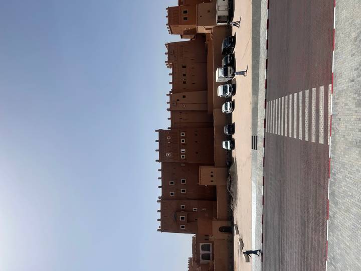A massive earthen-colored kasbah wall with parked vans lined along the base under a cloudless sky.