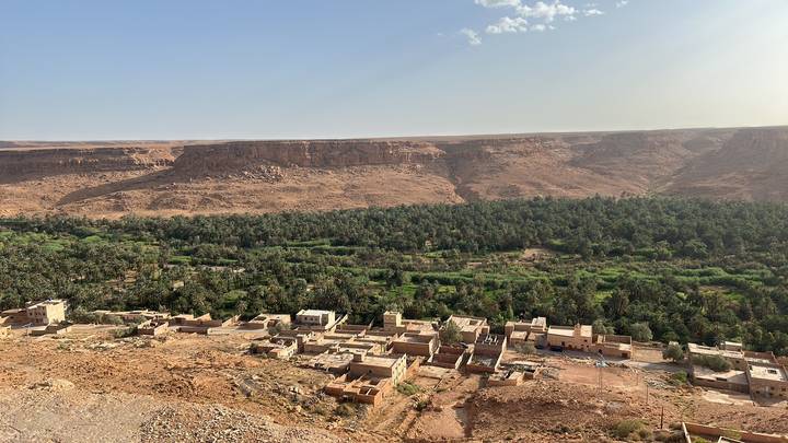 An elevated view of a lush palm oasis and small village set against arid desert cliffs.