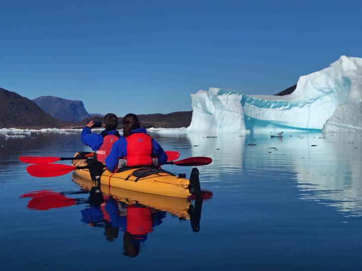 Two kayakers paddle past a towering iceberg reflected in calm Arctic waters.