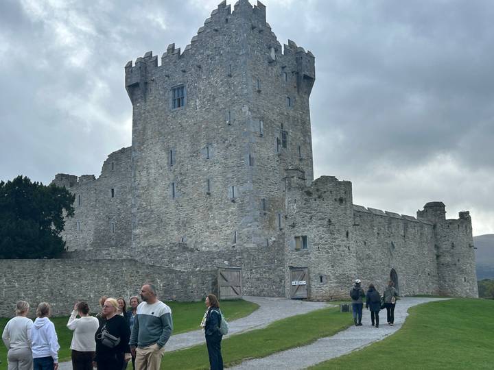 Stone castle tower under grey sky with tour group gathered on grassy courtyard.