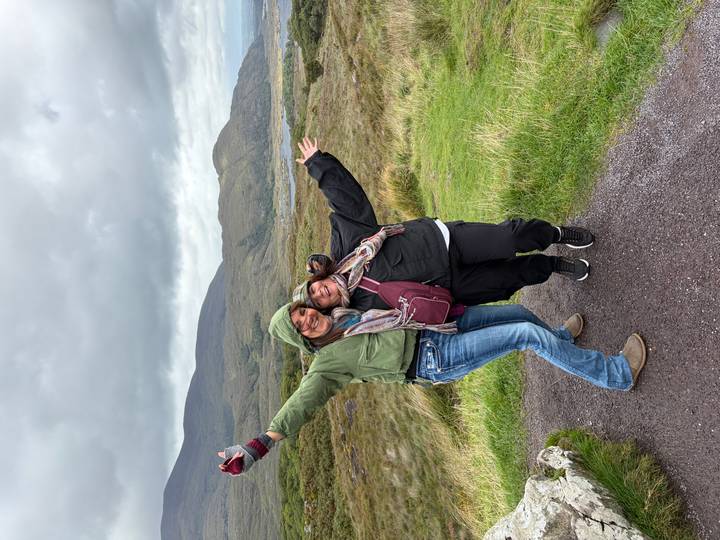 Two women in outdoor gear stretching arms wide on scenic hillside trail.