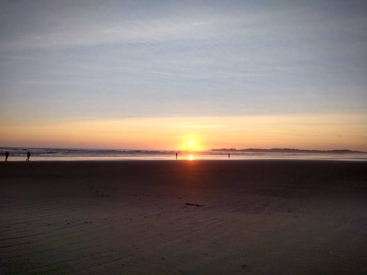 Sun sets over a wide sandy beach with a few silhouetted figures near the shoreline.