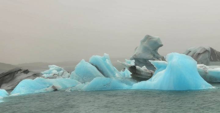 Blue icebergs float in a misty lagoon with streaks of black volcanic ash.