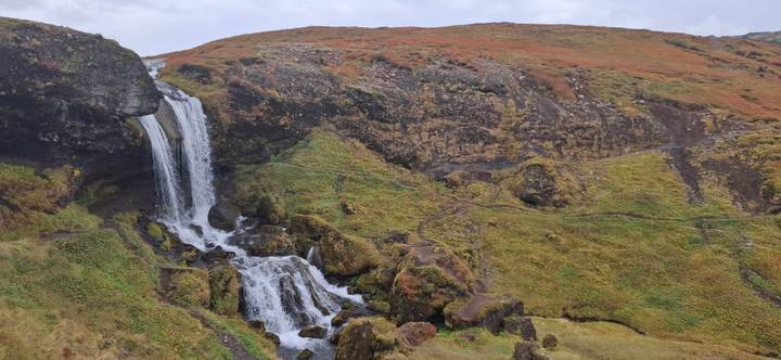 Waterfall cascades over dark rocks into a mossy ravine amid autumn-coloured hills.