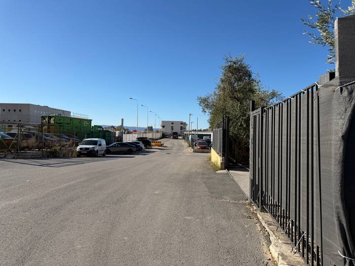 Industrial backstreet lined with parked cars, fencing and bright blue sky.