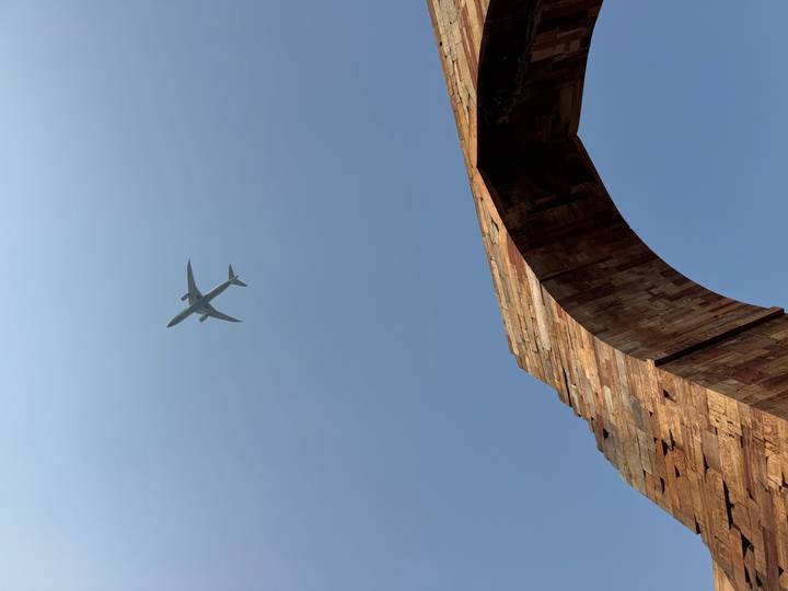 Airplane silhouetted against clear blue sky above an ancient stone arch.