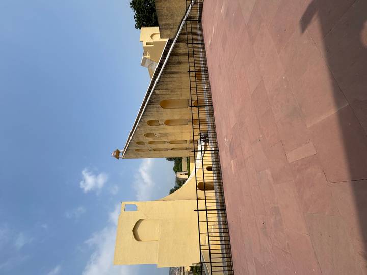 Large yellow sundial structure of Jantar Mantar under a clear blue sky with an open red-stone courtyard in front.