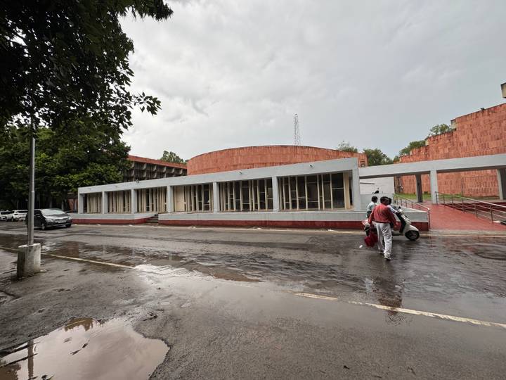 Rain-damp street outside a low modern auditorium as two travellers wheel luggage across wet pavement.