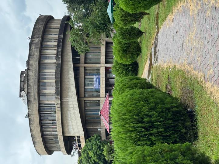 Circular modernist library building partially hidden behind manicured hedges on an overcast day.