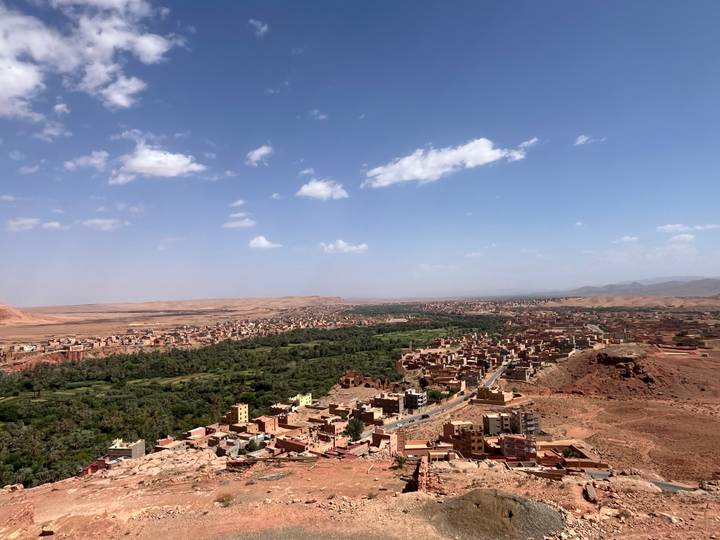 Sweeping aerial view over a green oasis valley surrounded by reddish desert hills and scattered villages.