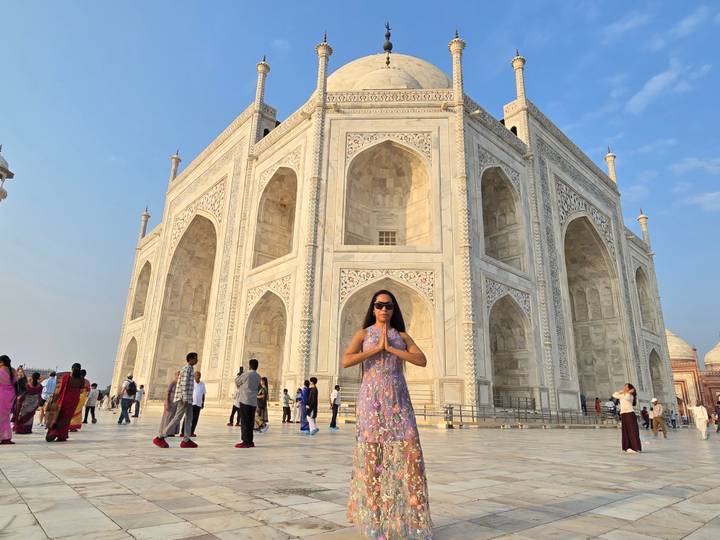 Woman posing with namaste gesture in front of the white marble Taj Mahal under a clear blue sky.