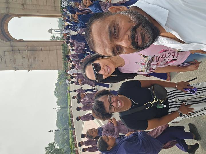 Three friends take a selfie at India Gate surrounded by school children and tourists.
