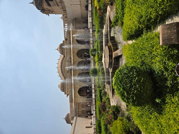 Fountain jets spray in a Mughal garden courtyard flanked by arched sandstone halls.