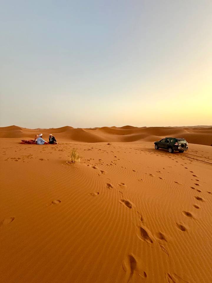 SUV and travelers rest atop golden dunes as the sun sets over the Sahara.