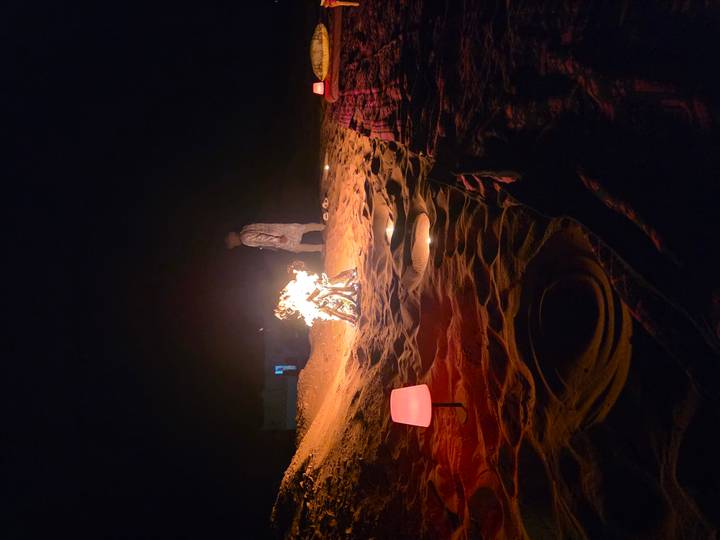 Nighttime desert campfire glows as a lone figure stands in the sand.