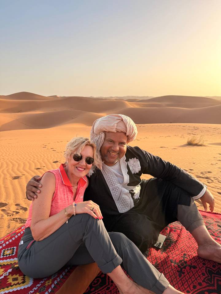 Smiling tourist and local guide pose together on golden desert sand at sunset.