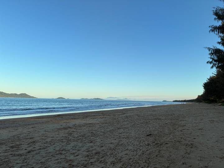 Empty sandy beach with calm blue sea and distant forested headlands under a clear sky.