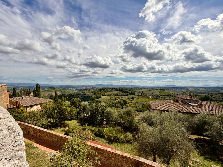 Expansive view over rolling Tuscan hills, vineyards, and scattered farmhouses under dramatic clouds.