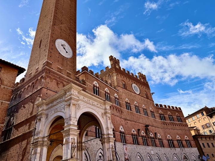 Historic brick tower with large clock rises against a bright blue sky in Siena’s main square.