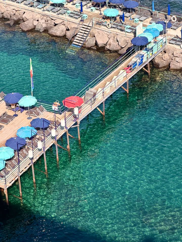 Pier with colorful umbrellas extends over sparkling turquoise water viewed from above.
