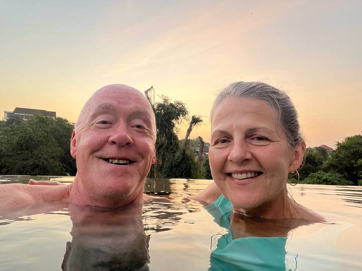 Smiling couple takes a relaxed selfie in an infinity pool at sunset with treetops behind.